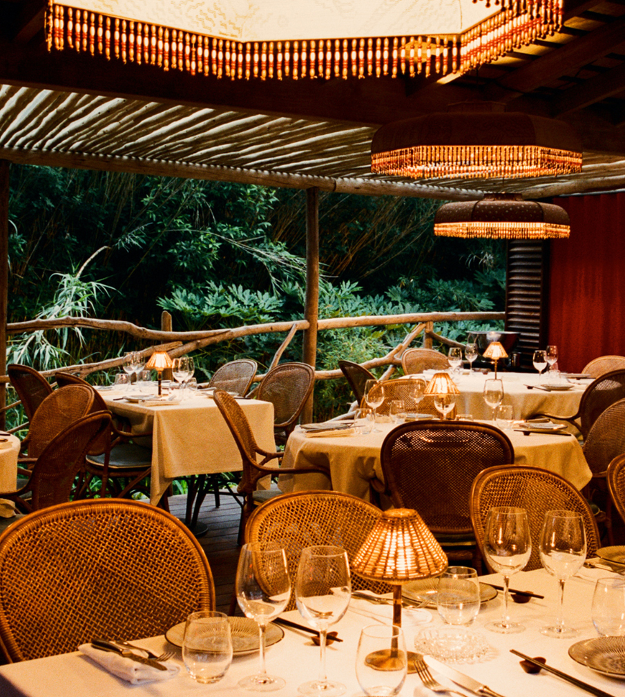 Tables dressées en rotin sous lampes suspendues à franges, terrasse couverte en bambou la nuit – restaurant La Sauvageonne, Ramatuelle