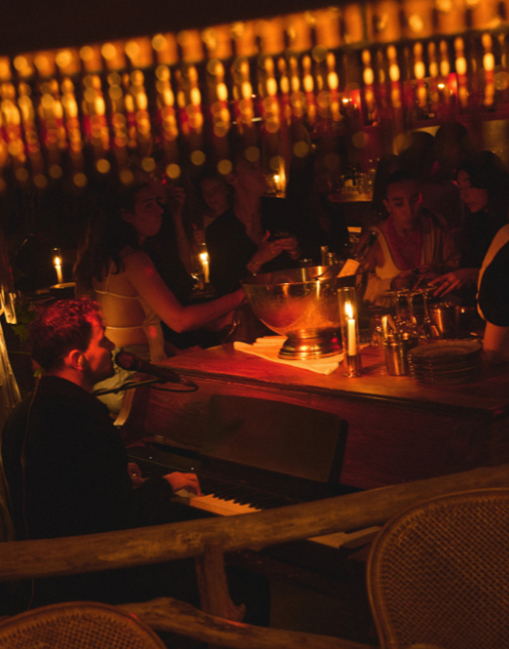 Lampe tressée et verres à vin sur table en bois entourée de feuilles tropicales, nuit – restaurant La Sauvageonne, Ramatuelle