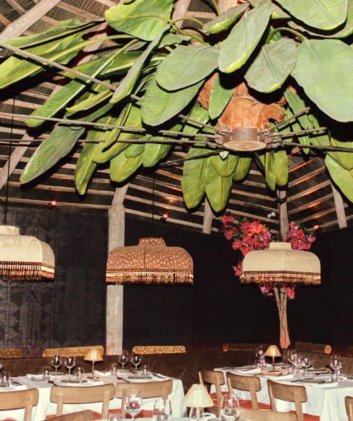 Table dressée en intérieur sombre avec lampe tressée et guirlande de bougainvilliers rouges, applique laiton au mur – restaurant La Sauvageonne, Ramatuelle