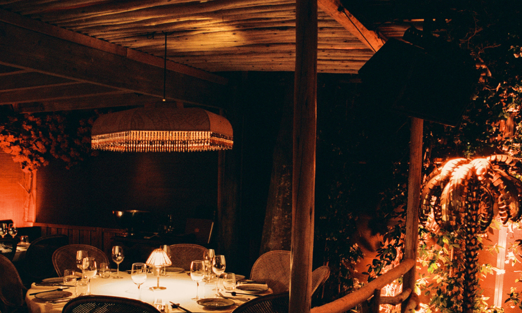 Table ovale en bois dress&eacute;e avec bougie et verres, palme dor&eacute;e et cloison de rondins blancs en arri&egrave;re-plan &ndash; restaurant La Sauvageonne, Ramatuelle